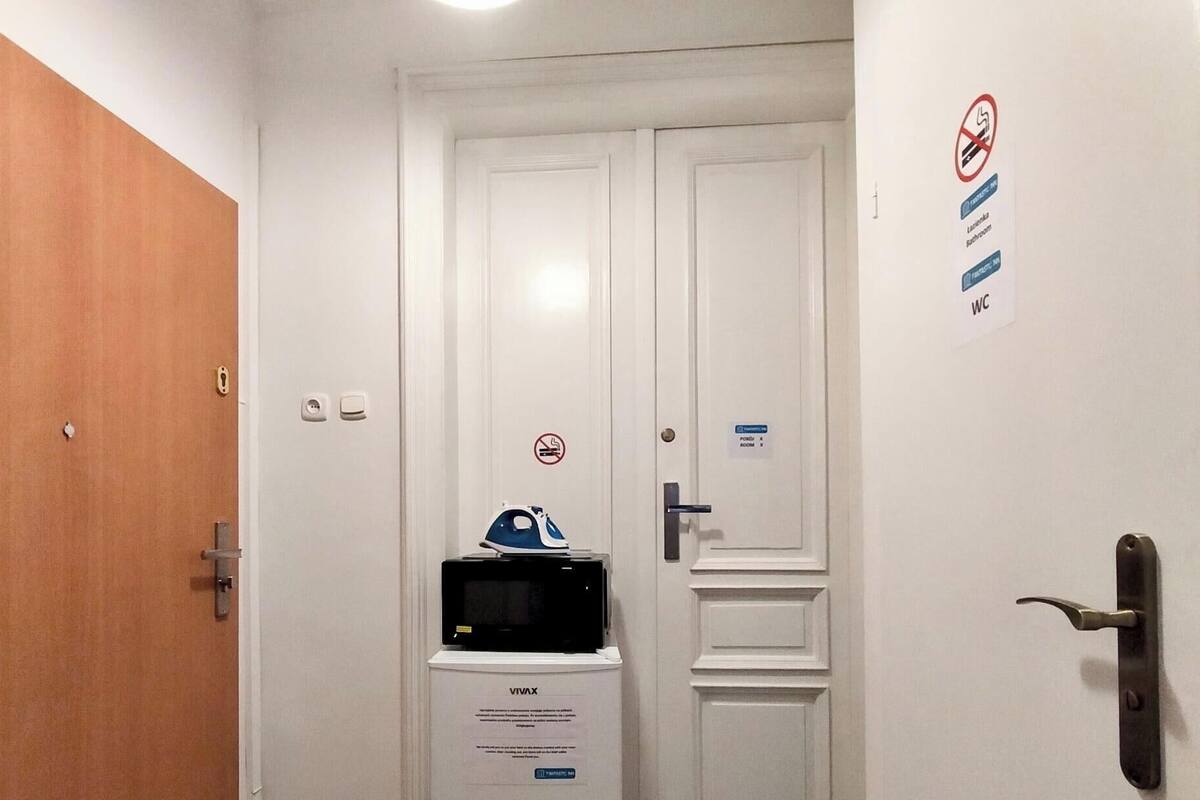 A hotel room with a wooden door, a white wall with a no-smoking sign, a microwave on a white stand, and a door with a keyhole.