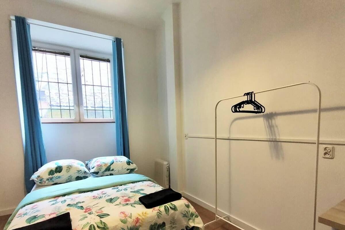 A bedroom with a bed, a window with blue curtains, and a metal drying rack against the wall.