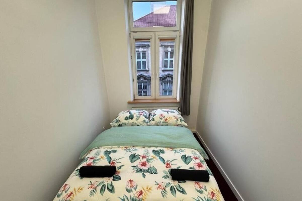 A narrow room with a bed, floral bedding, and a window with a view of buildings.