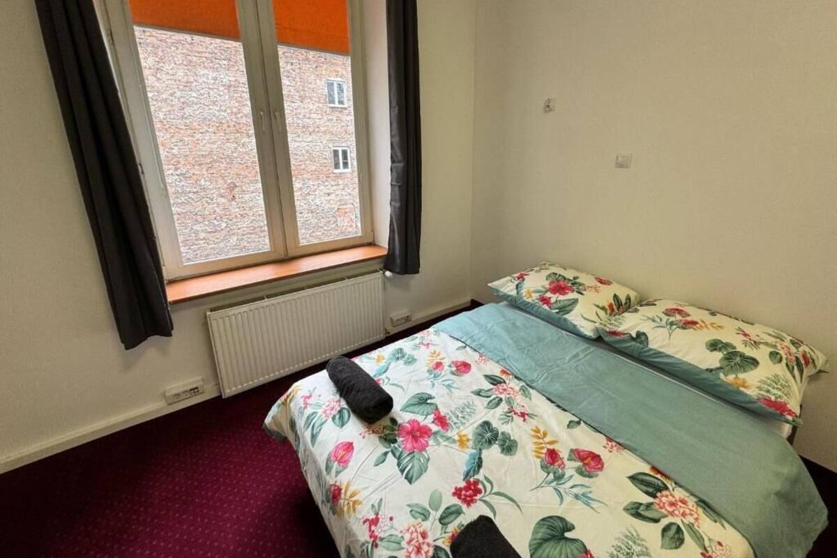 A bedroom with a bed, a window with curtains, a radiator, and a view of a brick building outside.