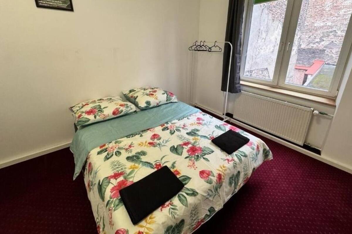 A small, single-bed room with floral bedding, a radiator, and a window with a view of a brick building.