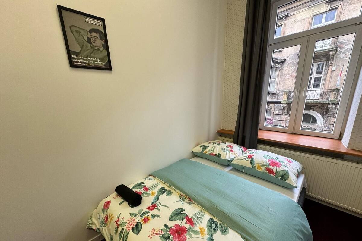 A bedroom with a bed, floral bedding, a window with a view of buildings, and a framed picture on the wall.