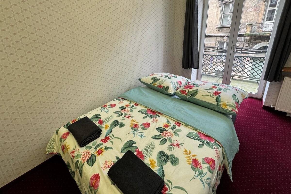 A bed with floral bedding and two pillows, a red carpet, and a window with a view of a building and a fence.