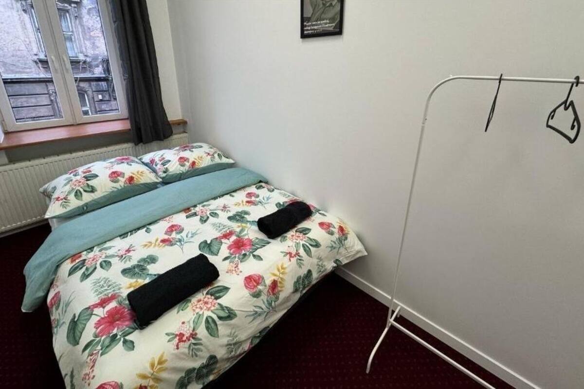 A bedroom with a floral bedspread, a window with a view of buildings, and a metal rack with a hanging item.