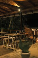 Terraza de madera al aire libre con mesas y bancos, rodeada de palmeras.