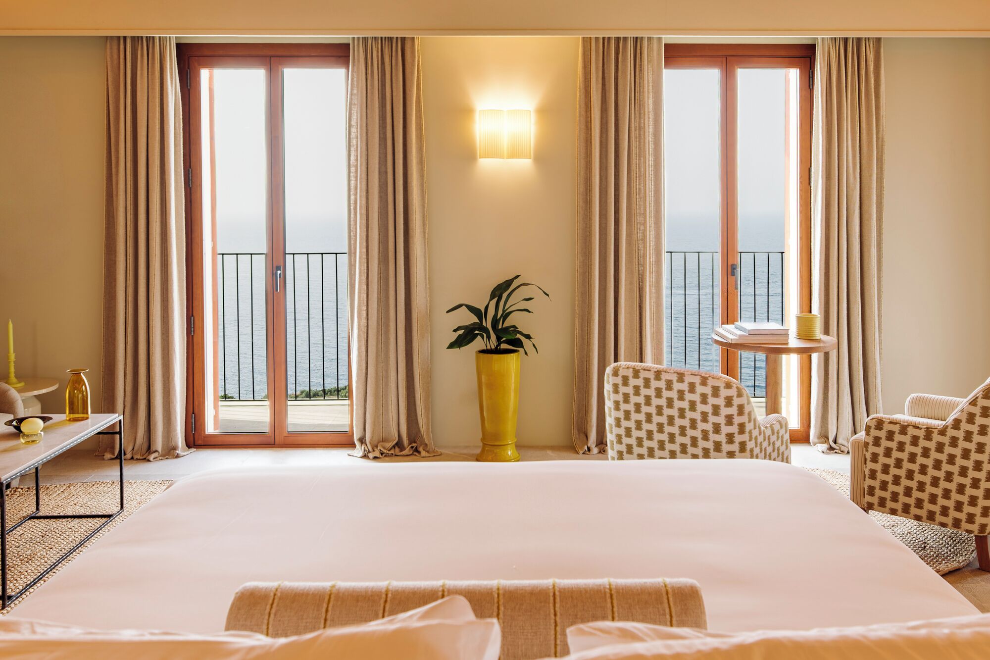 A hotel room with a large bed, two chairs, a small table, a potted plant, and a view of the sea through large windows.