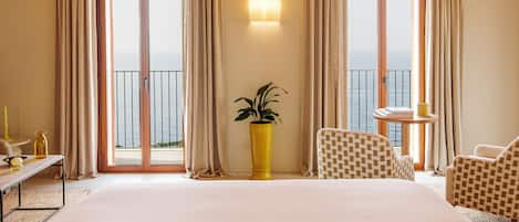 A hotel room with a large bed, two chairs, a small table, a potted plant, and a view of the sea through large windows.