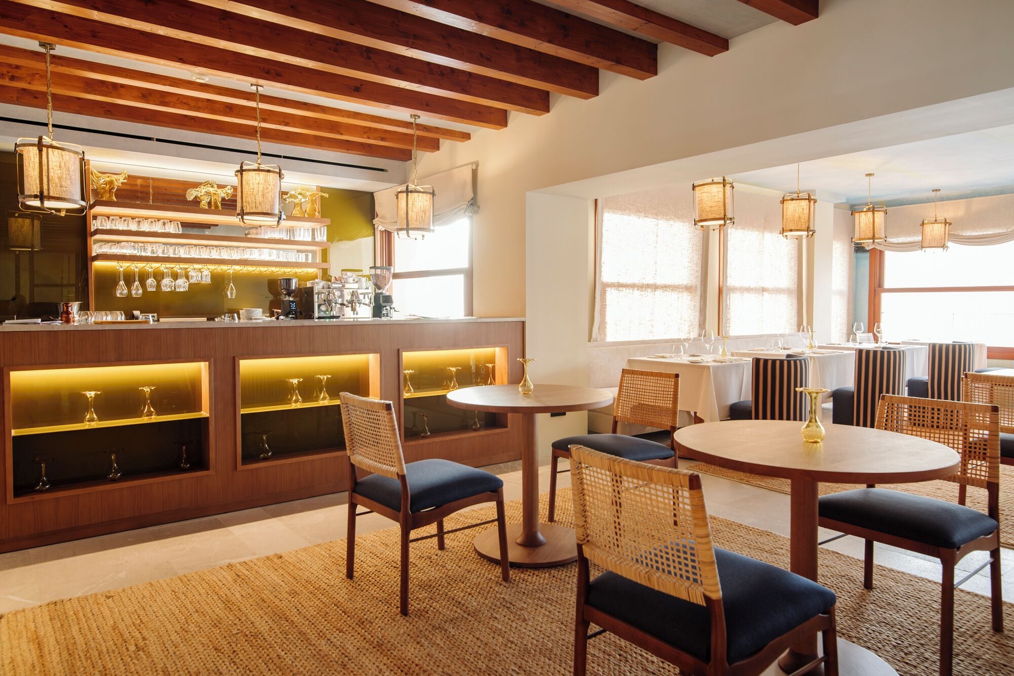 Un espace bar avec un plafond en bois et des luminaires suspendus, un comptoir avec des verres et une machine à café, ainsi que des sièges avec des tables rondes et des chaises en osier.