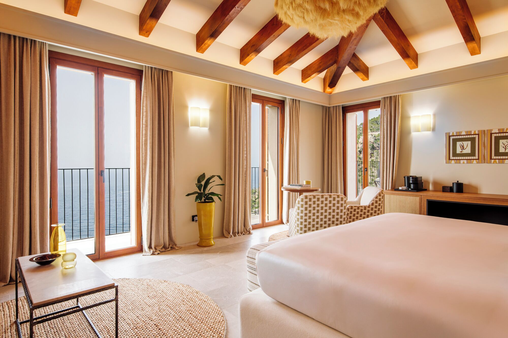 A hotel room with a large bed, a small table, a chair, a potted plant, and a balcony with curtains.