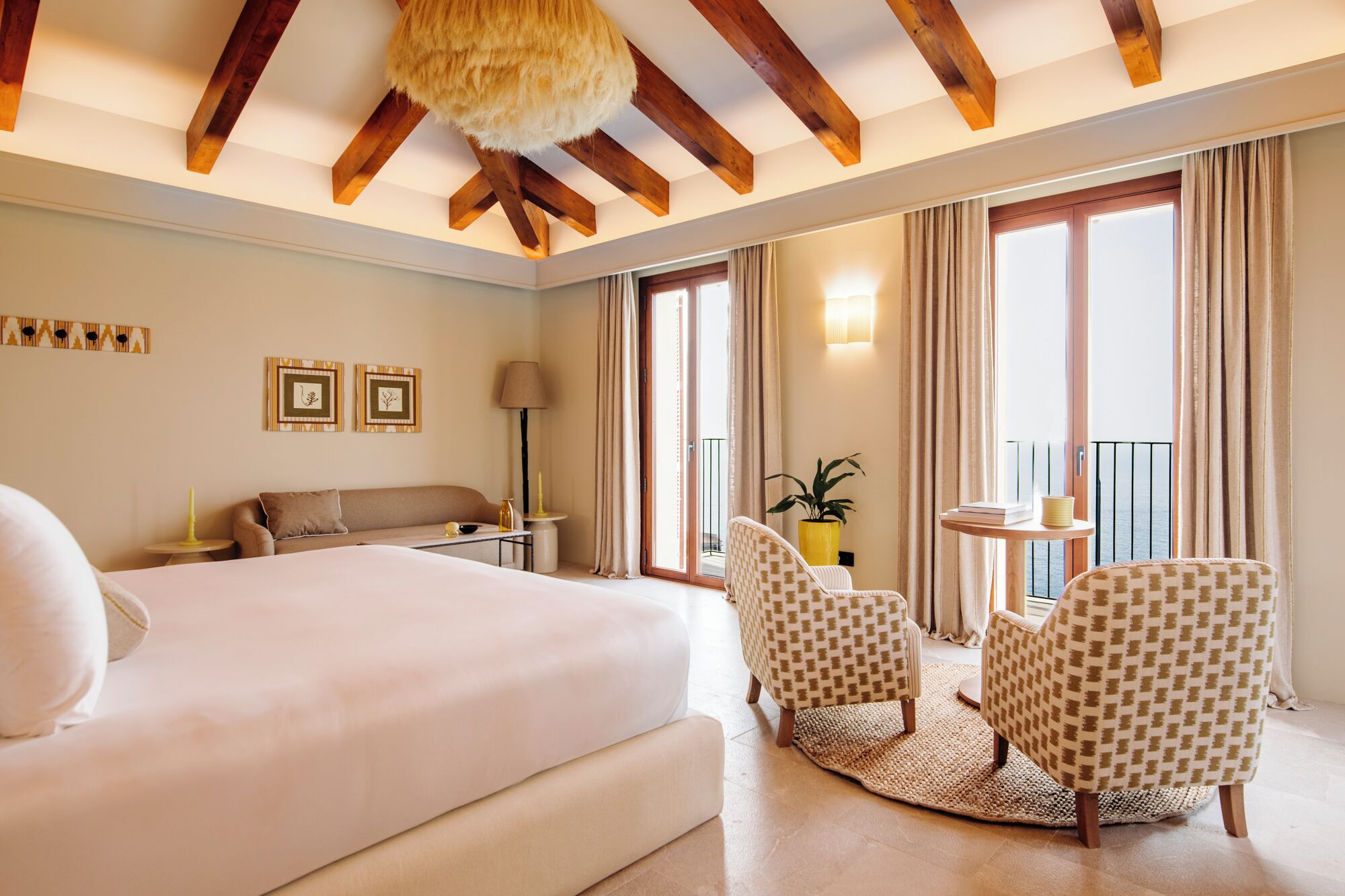 A hotel room with a bed, a sofa, two armchairs, a small table, and a balcony with curtains.