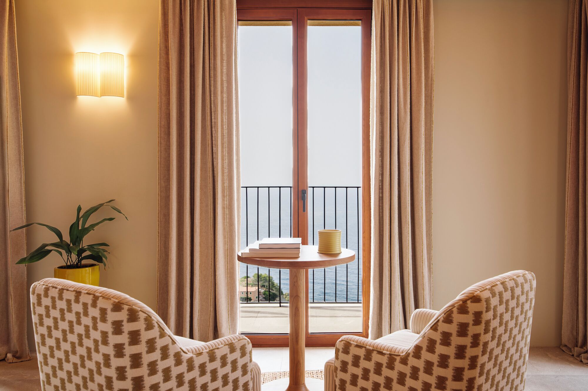 A room with two patterned chairs, a small round table with books and a yellow object, a potted plant, and a view through a large window with curtains.