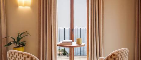 A room with two patterned chairs, a small round table with books and a yellow object, a potted plant, and a view through a large window with curtains.