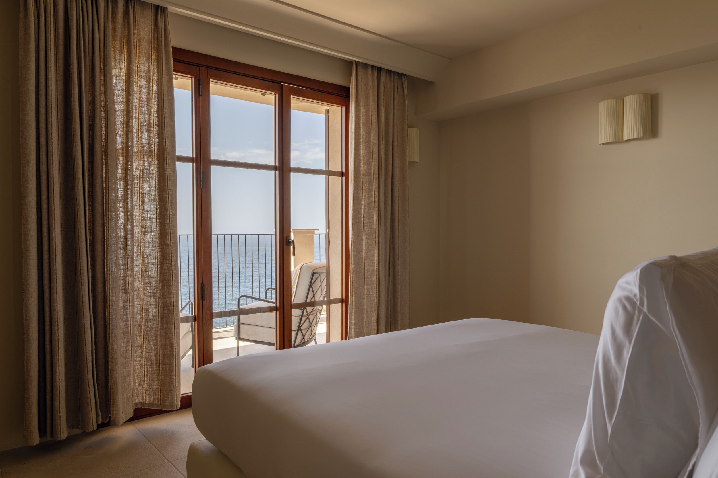 A hotel room with a large bed, a balcony with a chair, and a view of the sea.
