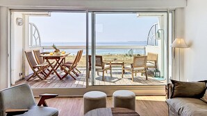 Een kamer met een grote schuifglazen deur die uitkomt op een terras met zitplaatsen en uitzicht op het strand.