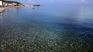 Vista costera con aguas transparentes y poco profundas que dejan ver guijarros y una costa lejana con edificios.