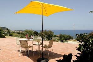 An outdoor patio with a table and chairs, a yellow umbrella, and a view of the sea. - Villa Bruca is a two-family house composed of two separate units.The unit offered, Villa Bruca 1, includes a double bedroom with the possibility of adding two single beds, a spacious bathroom with a modern walk-in shower, and a kitchen-living area equipp (Caronia)