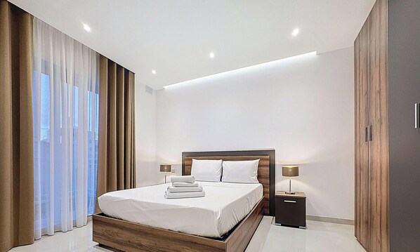 A modern bedroom with a wooden bed frame, white linens, and a nightstand with a lamp.