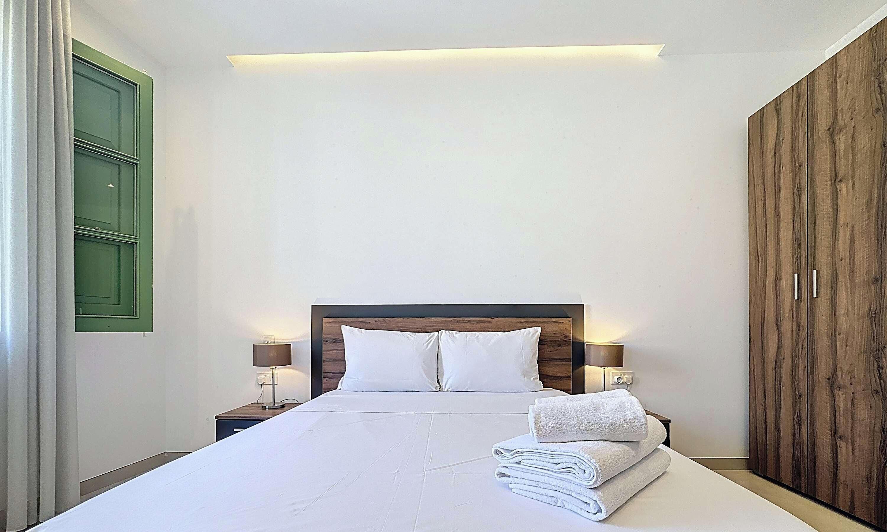 A neatly made bed with white linens and a folded towel, two bedside lamps, a wooden headboard, and a green door.