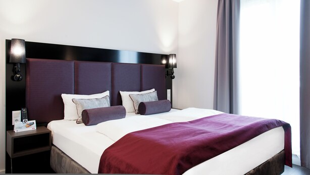 Ein Hotelzimmer mit einem großen Bett, einem Kopfteil mit integrierter Beleuchtung, einem Nachttisch mit Fernbedienung und einem Fenster mit Vorhängen.