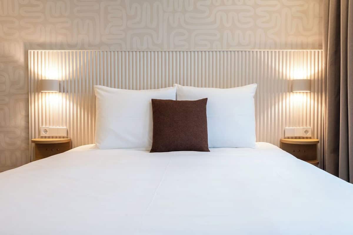 Cama bem arrumada com almofadas brancas e castanhas, duas luminárias de cabeceira e papel de parede estampado.