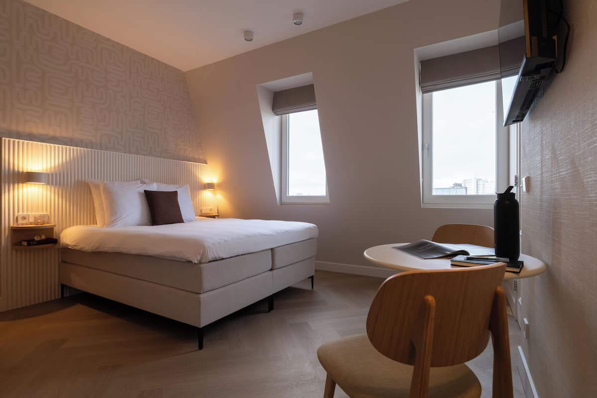 Quarto de hotel moderno com cama, abajures, uma cadeira, uma mesinha e uma janela com vista para a cidade.