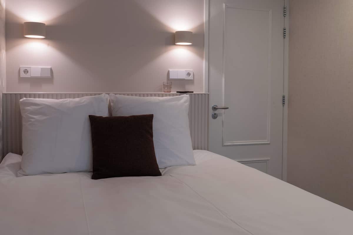 Quarto de hotel com cama, duas luminárias na parede, uma porta e um termostato.
