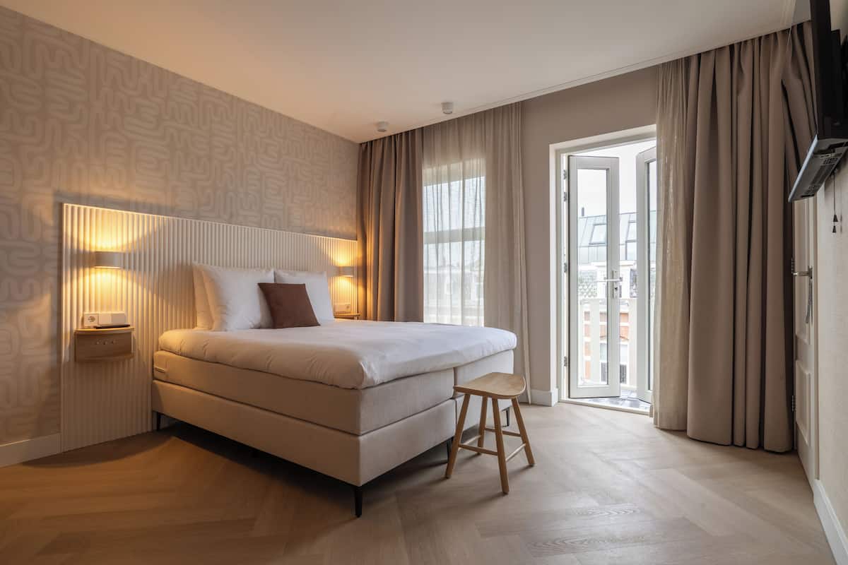 Quarto de hotel moderno com uma cama grande, luminárias laterais, um banquinho de madeira e vista através de uma porta de vidro.