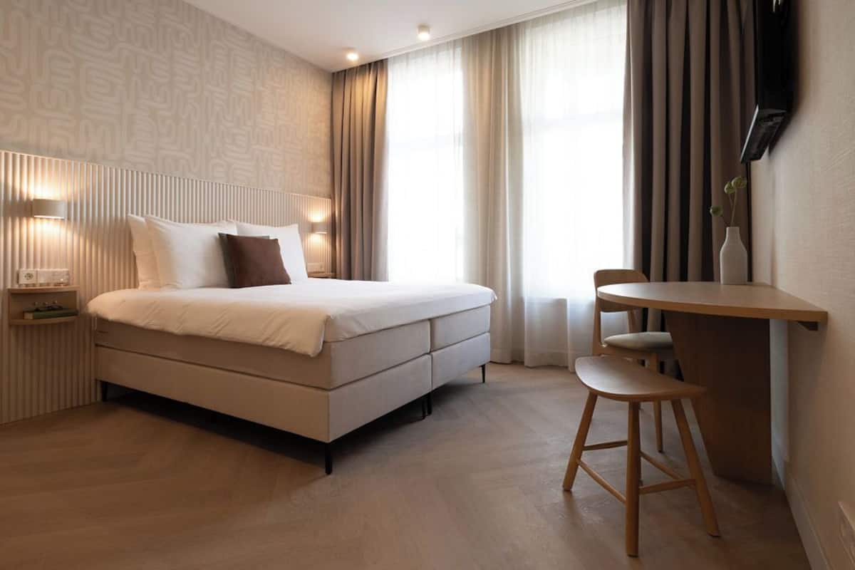 Quarto de hotel moderno com cama, mesa de cabeceira, luminária e uma pequena escrivaninha com cadeira.