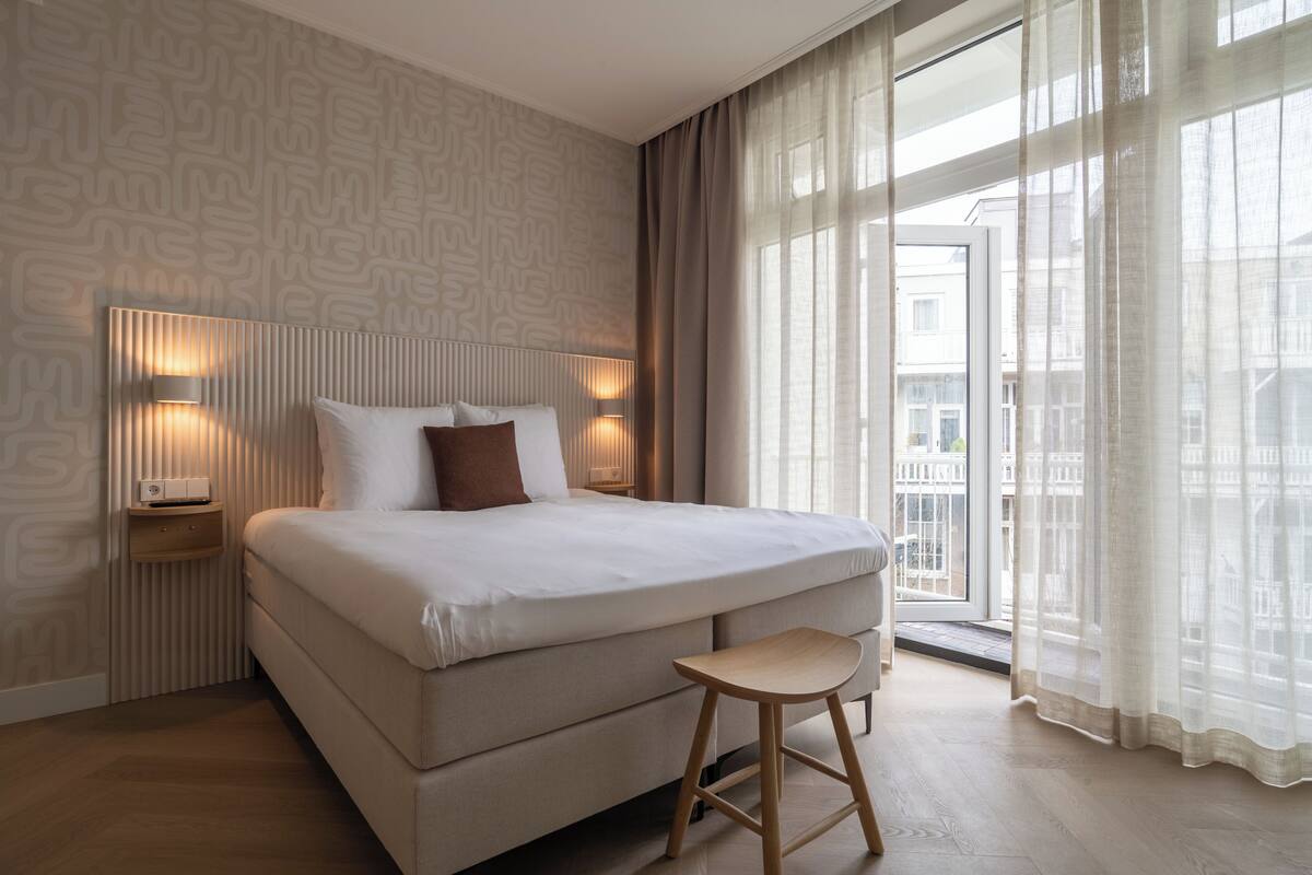Quarto de hotel moderno com uma cama grande, abajures, um banquinho de madeira e vista através de cortinas leves.