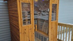 Una cabaña de madera con puerta y ventanas de vidrio, una terraza de madera y suelo cubierto de nieve.