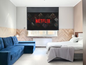 Une chambre d’hôtel moderne dotée d’un grand téléviseur à écran plat fixé au mur, sur lequel apparaît le logo de Netflix, d’un canapé bleu, d’un lit avec une couette blanche et d’un mur en bois avec un motif en chevrons.