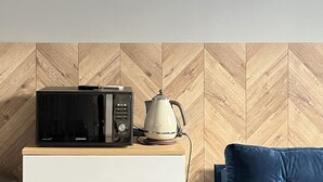 Eine Küchenzeile mit Mikrowelle und Wasserkocher auf einem Holzkasten, ein weißer Kühlschrank und ein blaues Sofa mit gelbem Kissen.