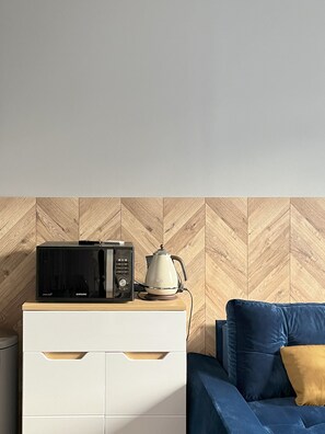 A kitchenette with a microwave and kettle on a wooden cabinet, a white refrigerator, and a blue sofa with a yellow pillow. - Comfy Apartment with projector, Jewish District, Rzeszowska (Kraków)