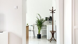 Un couloir avec une porte blanche sur la droite, un porte-manteau, une plante en pot et, au fond, un coin cuisine.