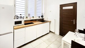 Cocina compacta con armarios blancos, encimera de madera, microondas, hervidor, y cafetera. Hay una ventana con persianas, una puerta, y un pequeño comedor con una copa de vino y un plato.