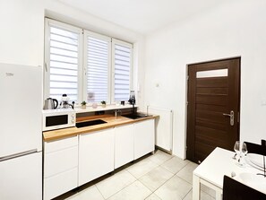 A compact kitchen with white cabinets, a wooden countertop, a microwave, a kettle, and a coffee machine. There's a window with blinds, a door, and a small dining table with a wine glass and plate. - Cozy Apartment for family next to Wawel II, Gertrudy 19 (Kraków)