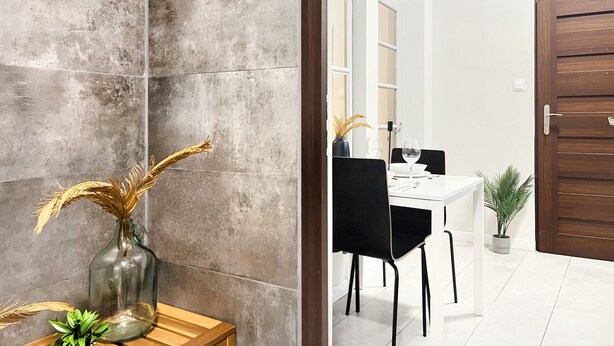 Un comedor moderno con una mesa blanca, sillas negras y un jarrón con plantas secas sobre una mesita de madera.