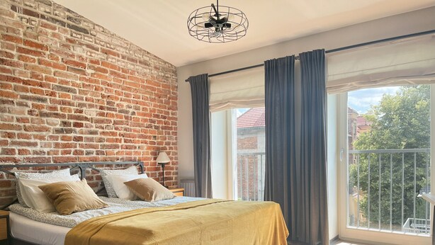 Un dormitorio con pared de ladrillo, cama con cubrecama amarillo, mesita de noche, lámpara y vistas al exterior desde un balcón.