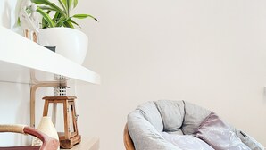 A room with a wicker chair, a white shelf with a potted plant, a decorative lamp, and a wall-mounted television.