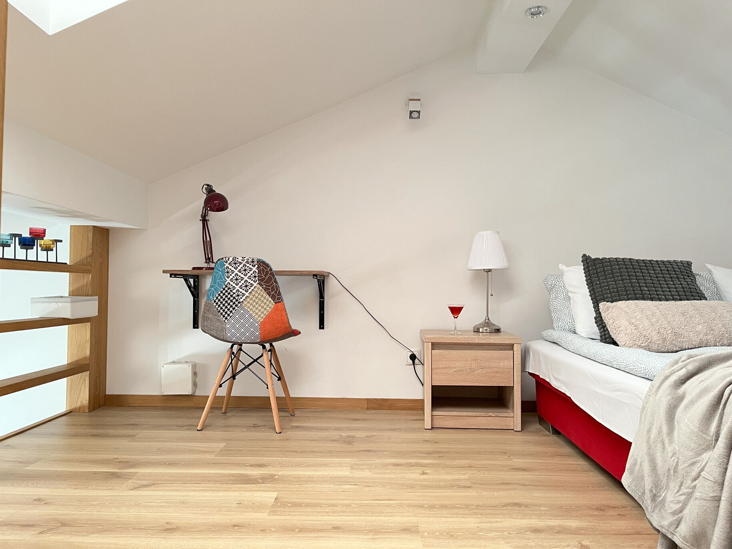 A bedroom with a bed, bedside table, lamp, chair, and a shelf with various items.