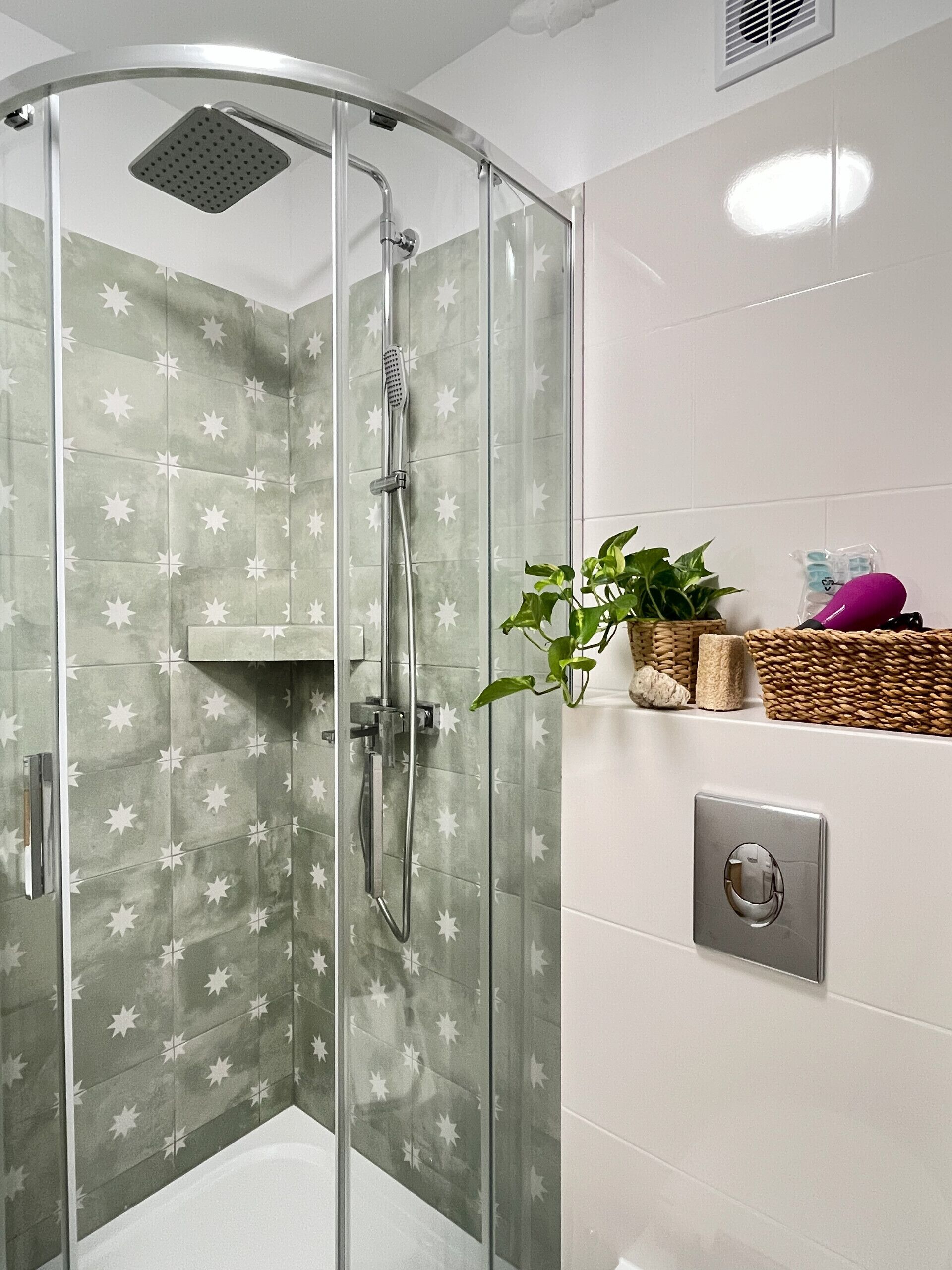 Ducha envuelta en vidrio con pared de azulejos con diseño de estrellas, una bañera blanca y un estante con una planta y artículos de aseo.