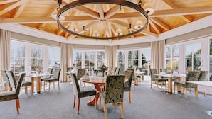 Un vaste espace dédié à la salle à manger, avec des tables rondes et des chaises à motifs, de grandes fenêtres et un plafond en bois.