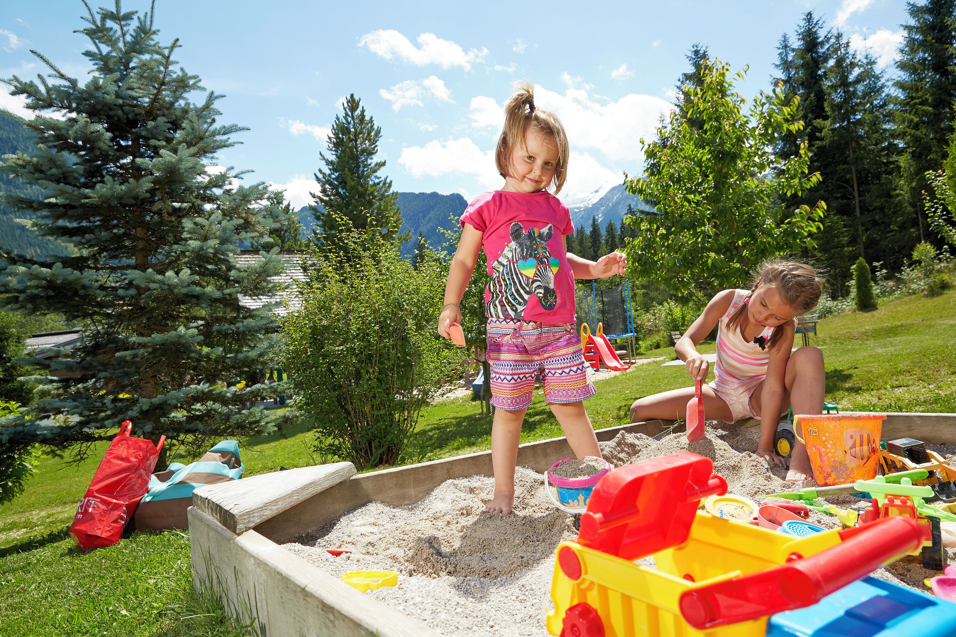 Children playing in a sandbox with toys and a red bag in a grassy outdoor area with trees and mountains in the background.