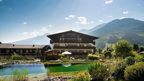 Un chalet de montagne avec un grand bâtiment, un étang et une végétation luxuriante tout autour.