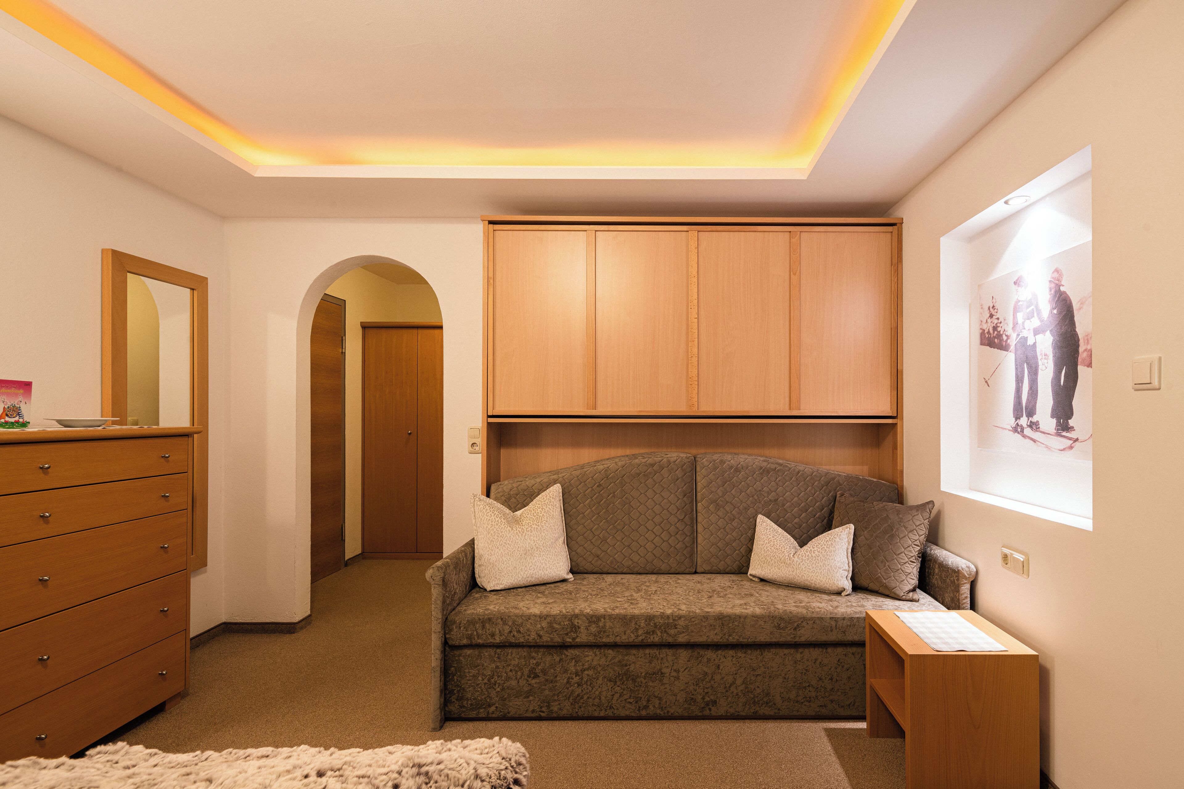 Ein Zimmer mit einem Holzkasten, einem grauen Sofa mit Kissen, einem Holzschrank über dem Sofa und einem gerahmten Bild an der Wand.