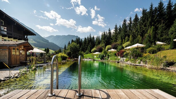 Une terrasse en bois avec une piscine, entourée d’une végétation luxuriante et de montagnes.