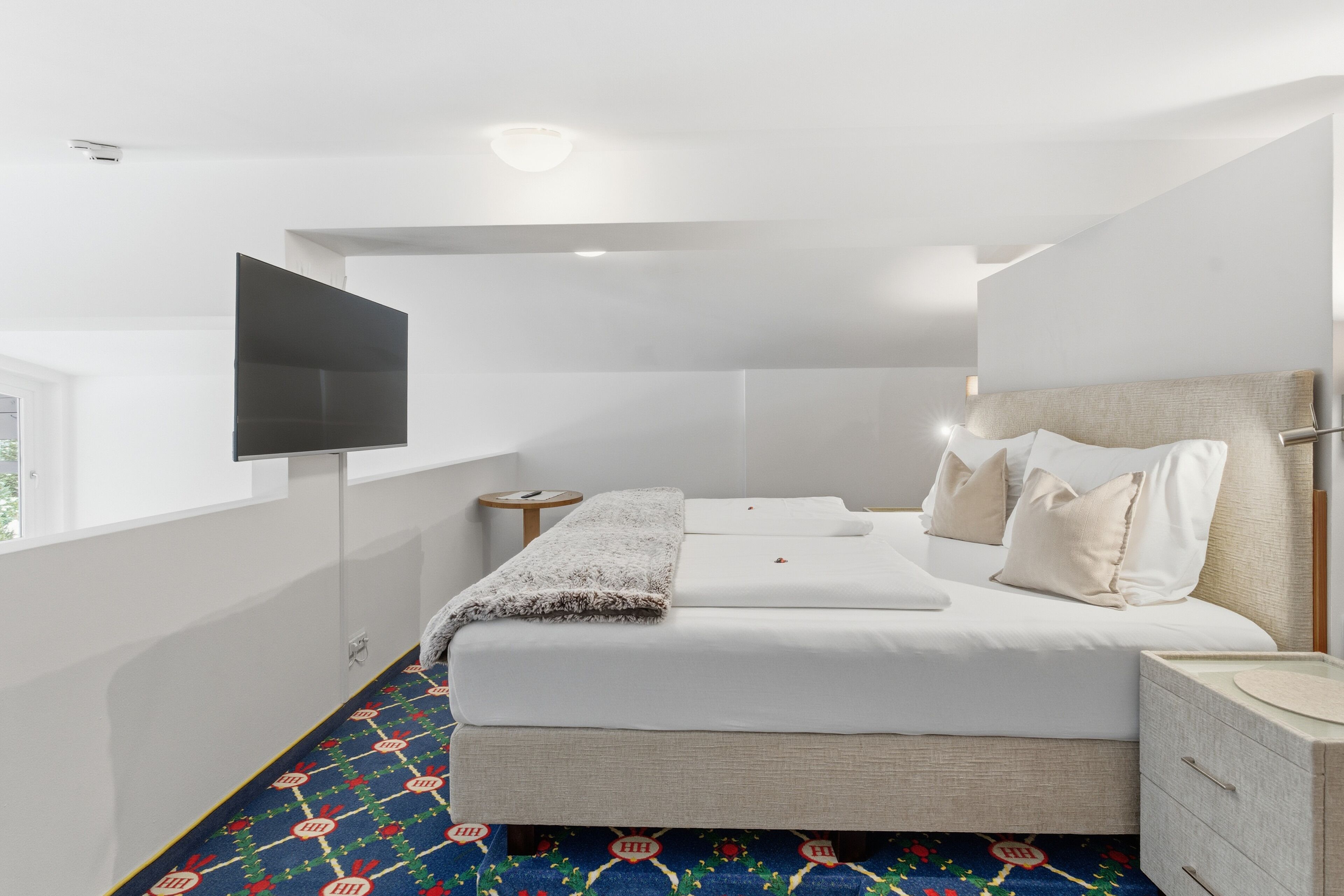 Une chambre à coucher moderne avec un lit, des tables de chevet, un téléviseur fixé au mur et un tapis à motifs.