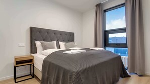 A modern bedroom with a tufted headboard, a large window with curtains, a wooden nightstand, and a neatly made bed. - EIR Three Bedroom Penthouse (Reykjavík)