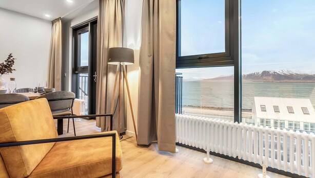 A modern hotel room with a large window offering a view of a body of water and mountains, a cozy seating area with a sofa and armchair, and a floor lamp. - EIR Three Bedroom Penthouse (Reykjavík)