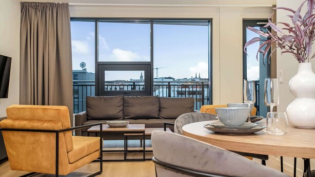 A modern living room with a sofa, armchairs, a dining table, and a view of a cityscape through large windows. - EIR Two Bedroom Apartments (Reykjavík)